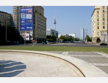 Straßenszene in einer Stadt mit einer breiten Straße, flankiert von mehrstöckigen Gebäuden, im Hintergrund der Berliner Fernsehturm. Im Vordergrund befindet sich eine niedrig geschwungene Steinmauer mit Grünflächen dahinter.