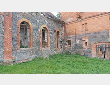 Verlassene Gebäudefassade aus Ziegelstein und Naturstein mit bogenförmigen Fenstern, teils ohne Glas, und überwuchertem Gras im Vordergrund. Rechts befindet sich ein Holztor, teilweise beschädigt, und die oberen Fenster sind mit Ziegelsteinen eingefasst.