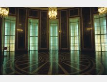 Großer, eleganter Saal mit hohen, bodentiefen Fenstern, die eine grünlich gefärbte Beleuchtung einlassen. Die marmorne Wandverkleidung, ein kunstvoll gemusterter Rundboden und große Kronleuchter dominieren den Raum.