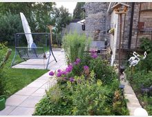 Gepflegter Hausgarten mit gepflastertem Weg und kleiner Terrasse mit Gartenschaukel und geschlossenem Sonnenschirm. Rechts steht ein Backsteinhaus mit Rankpflanzen, daneben ein dichtes Blumenbeet mit lila Blüten und Gartenfiguren am Rand.