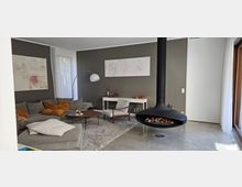 Wohnzimmer mit moderner Einrichtung, bestehend aus einer grauen Eckcouch mit orangefarbenen Kissen, einem niedrigen Holztisch, einem Teppich und einem hängenden schwarzen Kamin. Im Hintergrund eine graue Wand mit abstrakter Kunst, ein weißer Schreibtisch, ein Sessel und große Fenster mit weißen Vorhängen.