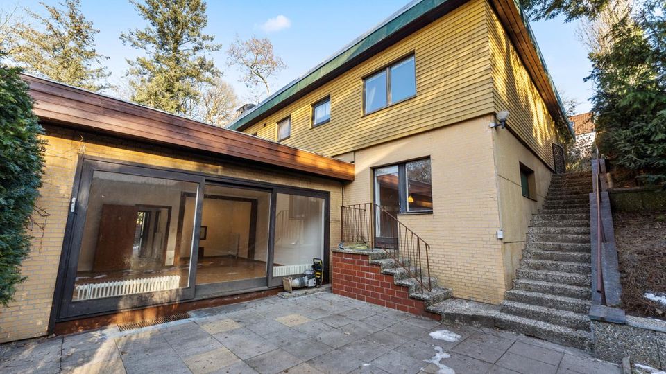 Innenhof eines zweigeschossigen Wohnhauses mit gelber Holzfassade und hellem Ziegel-Sockel, großer Glas-Schiebetürfront und gepflasterter Terrasse. Rechts führen Außentreppen mit Geländer am Hang entlang nach oben, umgeben von Bäumen und Sträuchern.