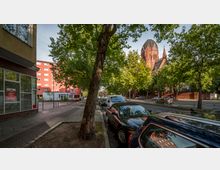Straßenszene mit parkenden Autos entlang eines baumgesäumten Gehwegs, einer roten Kirche mit einem auffälligen Turm und Kreuz im Hintergrund sowie modernen Wohngebäuden auf der linken Seite.