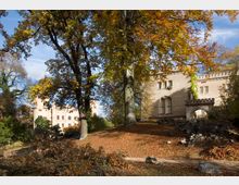 Schloss in einer herbstlichen Parklandschaft mit Laubbäumen und herabgefallenen Blättern. Das Gebäude hat sandfarbene Mauern, bogenförmige Fenster und Zinnen, daneben ein steinernes Torbogen-Element.