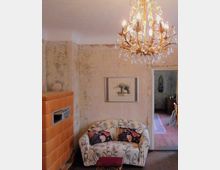 Ein Wohnzimmer mit einem kleinen, floralen Sofa, einem roten Hocker und einem Kaminofen aus orangebraunen Kacheln. Die Wände sind teils unverputzt, ein Kronleuchter mit Kristallanhängern hängt von der Decke, und ein gerahmtes Bild mit einem Elefanten ziert die Wand.