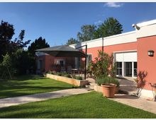 Ein einstöckiges, modernes Haus mit rosafarbenen Wänden und Flachdach, umgeben von einem gepflegten Garten mit grüner Rasenfläche und Pflanzkübeln. Eine Terrasse mit Sitzmöbeln und einem großen grauen Sonnenschirm liegt an der Hausfront.