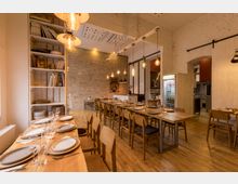 Ein warm beleuchteter Essbereich mit langen Holztischen, gedeckt mit Tellern und Gläsern, umgeben von einfachen Holzstühlen. Im Hintergrund sind freiliegende Ziegelwände, Regale mit Geschirr und Büchern sowie eine offene Küche sichtbar.