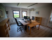 Ein kleines, modernes Büro mit einem Schreibtisch, zwei Bürostühlen und zwei Monitoren. Es hat helle Wände, Holzböden, drei Fenster mit Blick nach draußen und einfache Regale an den Wänden.