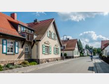 Straßenansicht einer ruhigen Wohngegend mit Einfamilienhäusern im traditionellen Stil, gekennzeichnet durch beige Fassaden, rote Satteldächer und Fensterläden in Grün und Weiß. Eine Person fährt mit dem Fahrrad auf der gepflasterten Straße, umgeben von kleinen Vorgärten und Laternen.