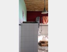 Badezimmer mit freiliegender Backsteindecke, weiß gefliesten Wänden und einer Dusche. Im Hintergrund befinden sich eine gemusterte Duschvorhangkombination, ein roter Vorhang sowie eine Holztreppe, die zur Duschwanne führt.