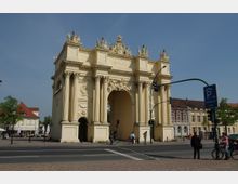Triumphalisches Tor auf einem öffentlichen Platz, verziert mit Säulen, Reliefs und Skulpturen, umgeben von historischer Architektur und einer Straße mit Fußgängern und Radfahrern; blauer Himmel im Hintergrund.