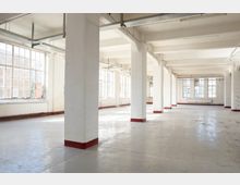 Großer, leerer Industrie- oder Loftinnenraum mit weißen Stützpfeilern, Betonboden und hoher Decke mit sichtbaren Leitungen und Trägern. Entlang der Außenwände befinden sich große, mehrteilige Fenster, durch die viel Tageslicht einfällt.