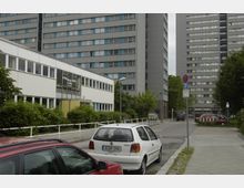 Straßenszene in einer urbanen Wohngegend mit mehreren Hochhäusern, einem niedrigen weißen Gebäude im Vordergrund und parkenden Autos entlang der Straße. Vegetation säumt den Gehweg, und mehrere Verkehrsschilder sind sichtbar.