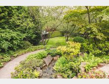 Ein Garten mit dichtem grünen Laub, einem kleinen Bachlauf und einem geschwungenen Gehweg, der zu einer Rasenfläche führt. Im Hintergrund befindet sich eine Hecke und eine kleine hölzerne Überdachung.