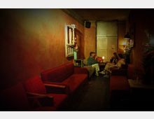 Dunkler Raum einer Bar oder eines Cafés mit rot-braunen Wänden und gemütlichen Polstermöbeln. Drei Personen sitzen an einem niedrigen Tisch, Kerzenlicht und Blumenarrangements sorgen für eine gedämpfte, intime Atmosphäre.