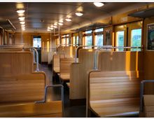 Innenraum eines historischen Zugwaggons mit Holzbänken und gelben Wandverkleidungen. Leuchtstofflampen sind an der abgerundeten Decke angebracht, und durch die Fenster fällt Tageslicht herein.