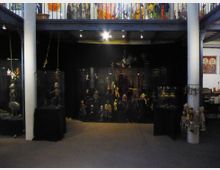 Ein Ausstellungsraum mit Marionetten und Puppen, die in beleuchteten Glaskästen präsentiert werden. Die Wände sind dunkel gestaltet, und darüber befindet sich eine Galerie mit bunt bemalten Figuren und Kunstwerken auf der Balustrade.