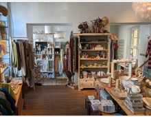Ein kleiner Geschäftsladen mit Holzfußboden, der Haushaltswaren, Dekorationen, Kleidung und Accessoires anbietet. Regale und Tische sind mit Schalen, Kerzen, Stoffen, Geschirr und anderen dekorativen Artikeln bestückt, während Kleidung auf Kleiderständern hängt.