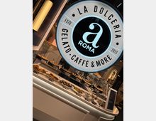 Ein modernes Café namens "La Dolceria" mit einem beleuchteten Rundlogo, das "Gelato, Caffè & More" zeigt. Im Vordergrund eine Vitrine mit einer Auswahl an Gebäck, Kuchen und anderen süßen Speisen.