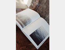 Ein aufgeschlagenes Buch liegt auf einem hölzernen Tisch und zeigt Fotografien von Wasseroberflächen. Im Hintergrund ist eine grob gemauerte Backsteinwand sichtbar.