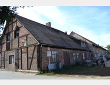 Ehemaliges Fachwerkhaus mit Backsteinfassade und schrägem Ziegeldach, erkennbar an verwitterten Balken und teils beschädigtem Mauerwerk, daneben gestapelte Baumstämme. Im Vordergrund ein Verkehrsschild mit Geschwindigkeitsbegrenzung auf 10 km/h.