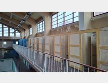 Ehemaliger Innenraum eines Schwimmbads mit Umkleidekabinen aus Holz, abgenutzten gelb-beigen Türen und abblätternder Farbe. Oberlichter mit roten Fensterrahmen lassen Tageslicht herein, und eine Metallbrüstung grenzt die obere Etage vom leeren Schwimmbecken ab.