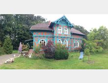 Fachwerkhaus mit roten Ziegeln und türkisfarbenen Holzverzierungen, umgeben von einem Garten mit Bäumen, Sträuchern und einer Liege auf einer Wiese; im Hintergrund befindet sich ein Wald.
