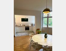 Helle Wohnküche mit Holzfußboden, einem runden Esstisch aus weißem Marmor und Stühlen im Vordergrund. Im Hintergrund eine offene Küche mit weißen Schränken, Fliesenspiegel und Einbaugeräten; rechts führt eine große Tür mit Fenster ins Freie.