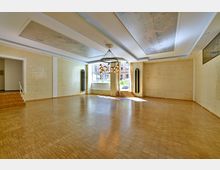 Großer, heller Raum mit Parkettboden, cremefarbenen Kachelwänden und modernem Kronleuchter aus Metall. Große Fenster mit Ausblick auf eine Terrasse und Treppenstufen an den Seiten verleihen dem Raum ein offenes Ambiente.