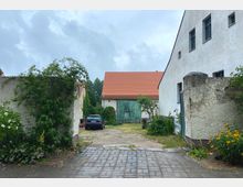 Hof mit gepflastertem Eingangsbereich, umgeben von alten verputzten Mauern und Gebäuden mit roten Ziegeldächern. Im Hintergrund steht eine Scheune mit großen, grünen Holztoren, während ein geparktes Auto und Pflanzen den Innenhof säumen.