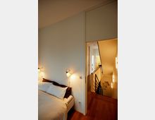 A modern interior showing a small bedroom with a white wall, a bed with neutral bedding, and wall-mounted reading lights. Adjacent to the bedroom is a hallway with wooden floors leading to a staircase with a black metal railing, illuminated by warm wall sconces.