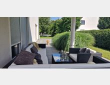 Moderne Terrasse mit Rattanmöbeln und weißen Polstern, flankiert von hohen Gräsern und umgeben von einem gepflegten Garten. Im Hintergrund ein Hund auf einem gepflasterten Weg, Bäume und ein Pool sichtbar.
