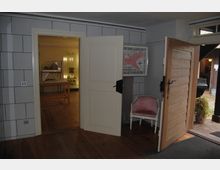 Blick in einen Eingangsbereich mit Holzboden und zwei geöffneten Türen, die jeweils in unterschiedliche Räume führen. Der linke Raum zeigt einen Ausstellungsbereich mit Holztischen und Vitrinen, während die rechte Tür nach draußen in einen gepflasterten Hof führt; ein rosa Stuhl steht an der Wand.