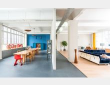 Großzügiger Open-Space-Innenraum mit zwei getrennten Bereichen: links ein Essbereich mit einem langen Holztisch, Holzstühlen und blauer Akzentwand, rechts ein Wohnbereich mit blauen Sofas, Holzboden und großen Fenstern mit gelben Vorhängen. Die Räume sind durch eine weiße Säule voneinander unterteilt.