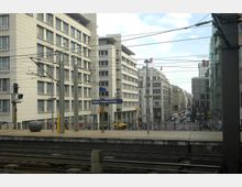 Bahnhofsumgebung in Berlin mit erhöhtem Bahnsteig, Gleisen im Vordergrund und modernen mehrstöckigen Gebäuden im Hintergrund. Ein Schild mit der Aufschrift „Berlin Friedrichstraße“ ist sichtbar, links und rechts verlaufen Straßen durch ein Geschäftsviertel.