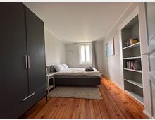 Schlicht eingerichtetes Schlafzimmer mit einem Doppelbett auf einem Teppich, einer Kommode links, einer weißen Regalwand rechts und einem schwarzen Kleiderschrank im Vordergrund. Das Zimmer hat Holzboden, weiße Wände, ein zentrales Fenster mit grauem Vorhang und ein Bild an der rechten Wand.