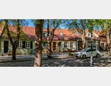 Tree-lined cobblestone residential street with a row of small pastel houses featuring red tiled roofs, shuttered windows, and front doors facing the sidewalk. Potted plants and benches sit along the facades, and a small white car is parked at the curb under leafy trees.