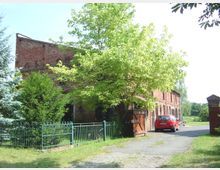 Backsteingebäude mit mehreren Bogenfenstern und einem asphaltierten Zufahrtsweg, flankiert von Bäumen und einem grünen Metallzaun. Rechts steht ein rotes Auto vor einer hölzernen Toröffnung.