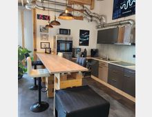 Moderne Gemeinschaftsküche mit rustikalem Holztisch, hohen Barhockern und grauen Einbauschränken. An der Wand sind Geräte wie ein Backofen, Mikrowelle und Kaffeemaschine integriert, während sichtbare Lüftungsrohre und Hängelampen im Industrial-Stil die Einrichtung ergänzen.