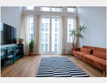 Wohnzimmer mit einem großen Fenster, das viel Tageslicht hereinlässt. Im Raum befinden sich eine orangefarbene Couch, eine schwarz-weiß gestreifte Teppichmatte, ein grünes Regal mit Fernseher und grüne Topfpflanzen.
