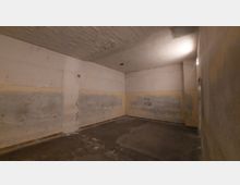 Leerer Raum mit unverputzten, teilweise verfärbten Wänden und Betonboden. Die Decke ist simpel und weist Risse auf, ein einzelnes Licht leuchtet den Raum schwach aus.