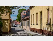Schmale, gepflasterte Gasse mit gelb gestrichenen Gebäuden auf der rechten Seite, roten Ziegeldächern und Fensterläden. Im Hintergrund sind weitere Häuser und ein geparkter Wagen sichtbar, umgeben von Bäumen.