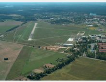 Luftaufnahme eines kleinen Flugplatzes mit einer Start- und Landebahn aus Asphalt, umgeben von Grünflächen und landwirtschaftlichem Gelände. Im Hintergrund sind Gebäude einer Ortschaft sowie Wälder und einzelne Seen zu sehen.