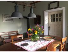 Gemütlicher Essbereich in einem rustikalen Raum mit Holztisch, Holzbänken und -stühlen, dekoriert mit frischen Blumen in einer Glasvase auf einer Tischdecke. Im Hintergrund weiße Tür mit Glasfenstern, ein Kaminrohr und eine Wand mit Schriftbild sowie zwei hängende Metalllampen.