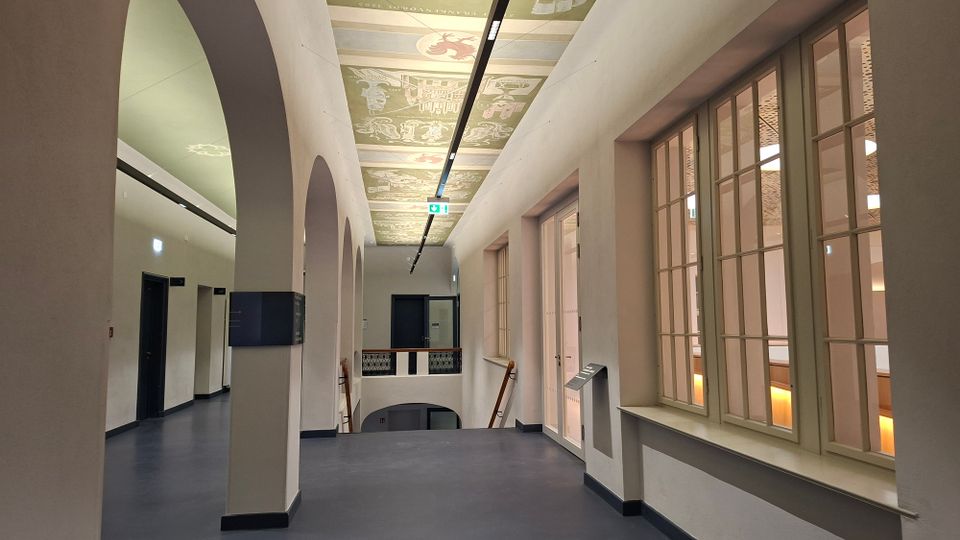 Heller Innenflur eines historischen Gebäudes mit hohen Rundbögen, großen Sprossenfenstern und einem zentralen Treppenabgang mit Geländer. An der Decke verläuft eine lange Lichtschiene unter bemalten Kassettfeldern mit dekorativen Motiven.