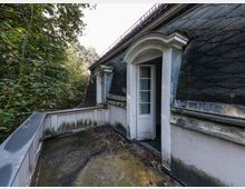 Kleiner Dachbalkon an einem älteren Gebäude mit dunkel gedecktem Mansarddach und Rundbogen-Dachgaube mit geöffnetem Türfenster. Der Betonboden und die Brüstung sind verwittert und von Moosspuren bedeckt, umgeben von dichtem Wald.