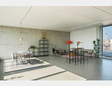 Modernes, minimalistisch eingerichtetes Loft mit Betonwänden, großem Fenster und Zugang zu einem Balkon mit Blick auf einen Fernsehturm. Der Raum umfasst einen Esstisch mit Stühlen, eine Stehtischgruppe, Regale, Pflanzen sowie dekorative Elemente wie eine Stehlampe und Trockenblumen.