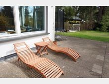 Zwei hölzerne Liegestühle und ein passender kleiner Tisch stehen auf einer gepflasterten Terrasse vor einem weißen Gebäude mit großen Fenstern. Im Hintergrund befinden sich ein Trampolin, ein Fußballtor und eine Rasenfläche, umgeben von Bäumen und einem Holzzaun.