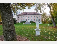 Herrenhaus in hellem Rosa mit symmetrischer Fassade, großen Fenstern und einem Ziegeldach, umgeben von einer Rasenfläche und Herbstbäumen. Im Vordergrund steht eine weiße, klassische Statue auf einem Sockel.
