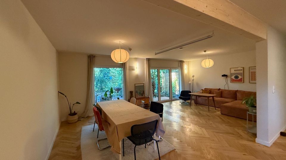 Ein modernes Wohnzimmer mit hellen Holzböden, einer großen Sitzecke und einem angrenzenden Essbereich mit einem Esstisch und Stühlen. Die rückseitige Wand besteht aus bodentiefen Fenstertüren, die auf einen Garten mit viel Grün blicken.